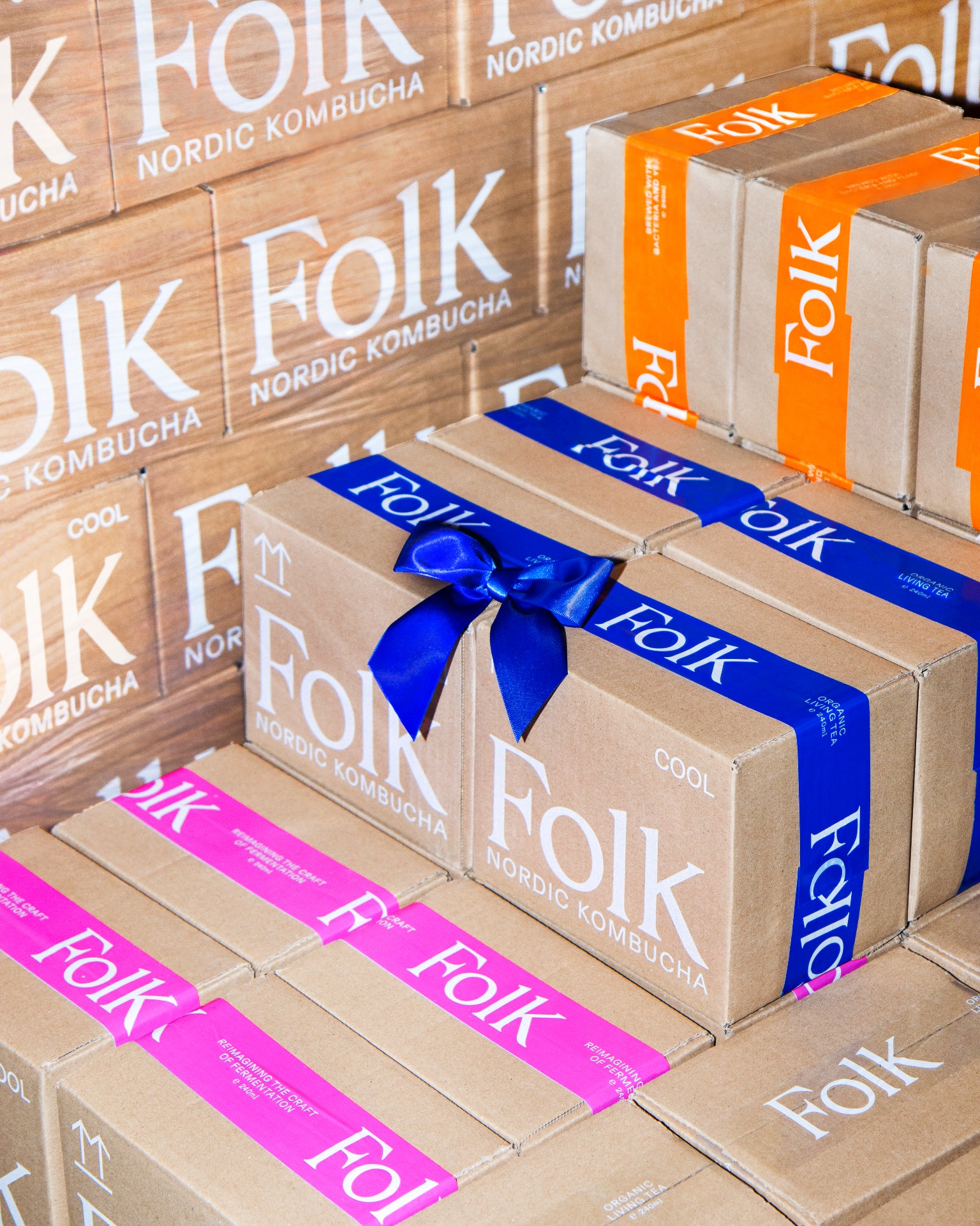 Stacked Folk Kombucha gift boxes with pink, blue and orange bands and a blue ribbon